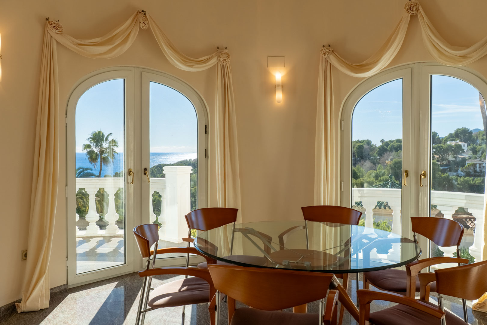 Eethoek met een glazen tafel en houten stoelen, voorzien van boogdeuren die toegang bieden tot het terras met balustrades en uitzicht op zee.