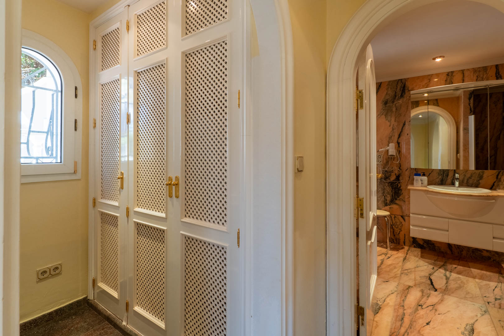 Gang met witte inbouwkasten met vlechtwerk en een boogingang naar een badkamer afgewerkt in roze geaderd marmer met een wit wastafelmeubel.