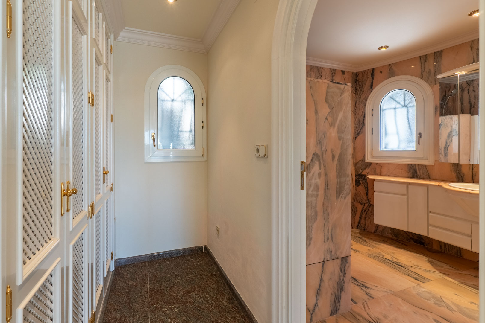 Gang met witte inbouwkasten met vlechtwerk en gouden beslag, die toegang biedt tot een badkamer volledig bekleed met roze marmer en boogvensters.