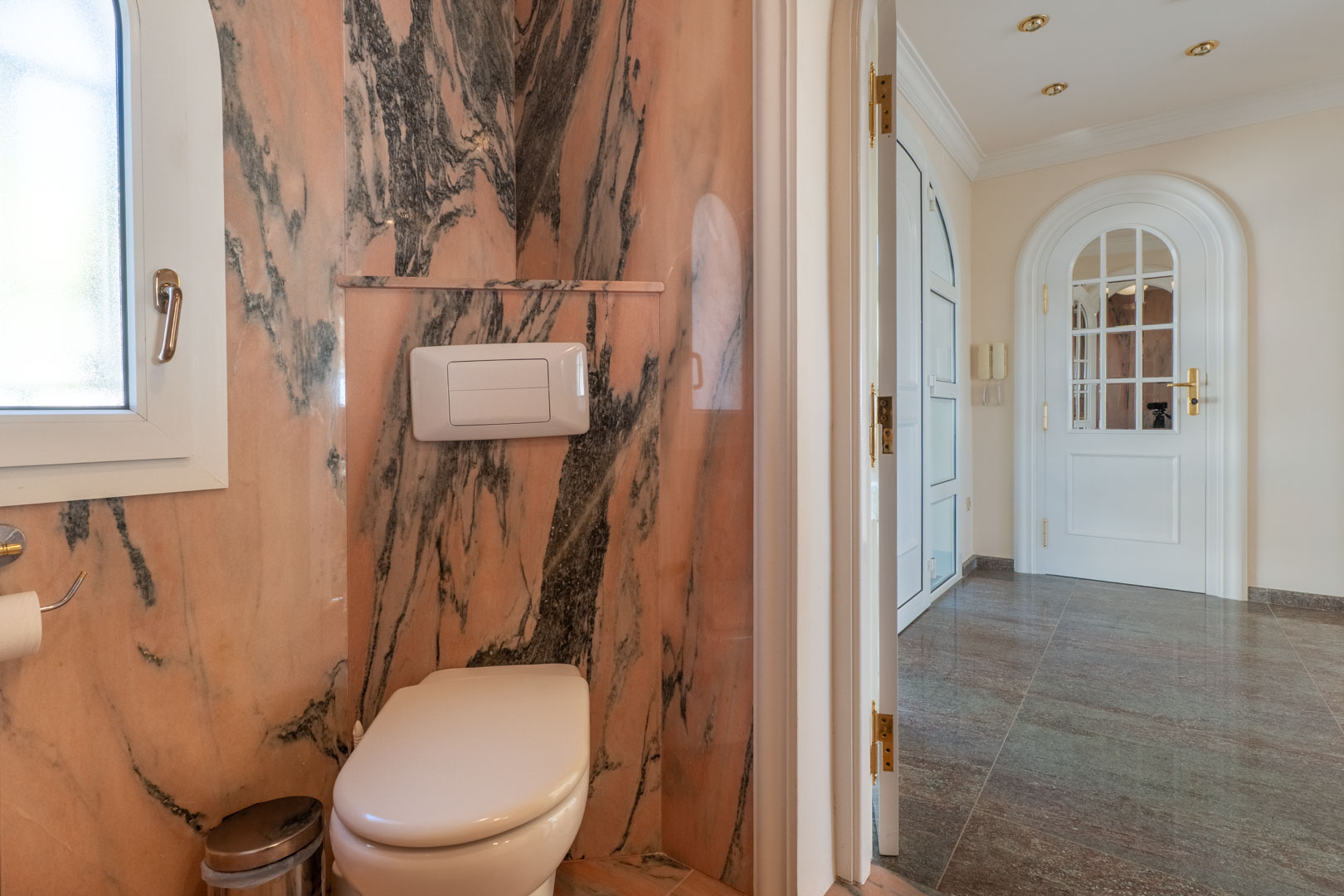 Gastentoilet met wanden van roze geaderd marmer, een hangend toilet en uitzicht op een gang met gepolijste stenen vloeren en witte boogdeuren.
