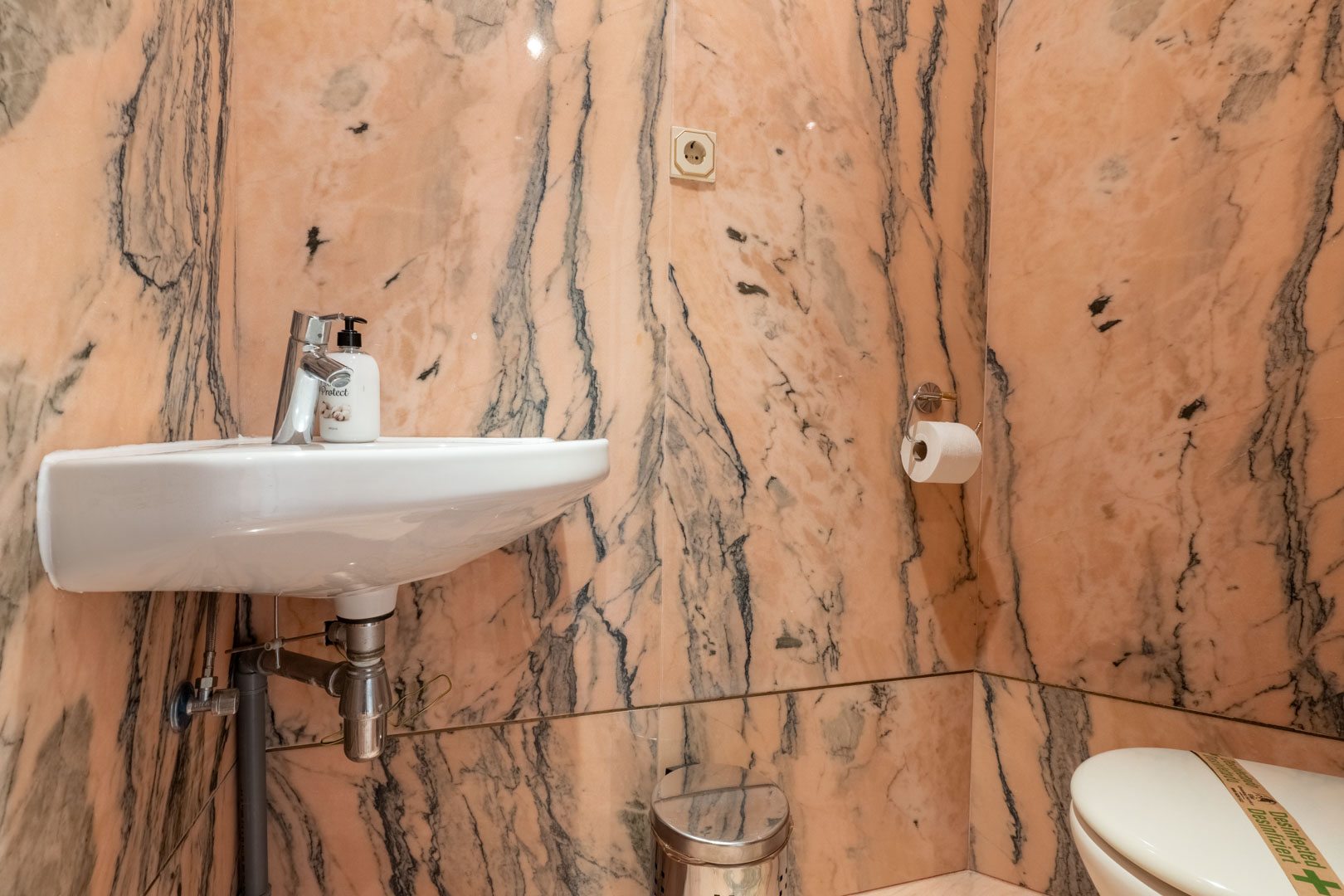 Gastentoilet volledig bekleed met roze marmer met donkere aders, voorzien van een wit hangend wastafelmeubel en chromen kranen.