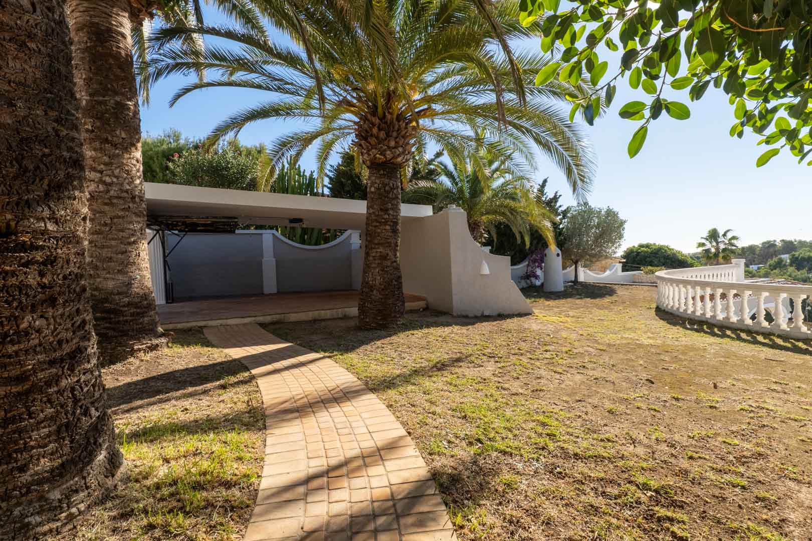 Geplaveid pad dat leidt naar een overdekte carport, omringd door volwassen palmbomen en een witte balustrade met vrij uitzicht.