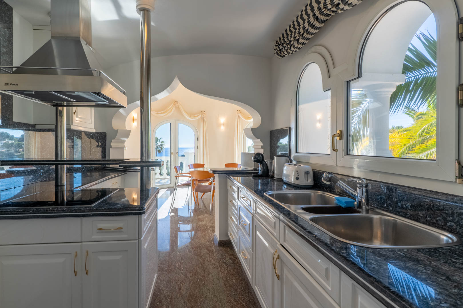Keuken met granieten werkblad en witte kasten, verbonden via een decoratieve boog met een lichte eetkamer die uitkijkt over de Middellandse Zee.