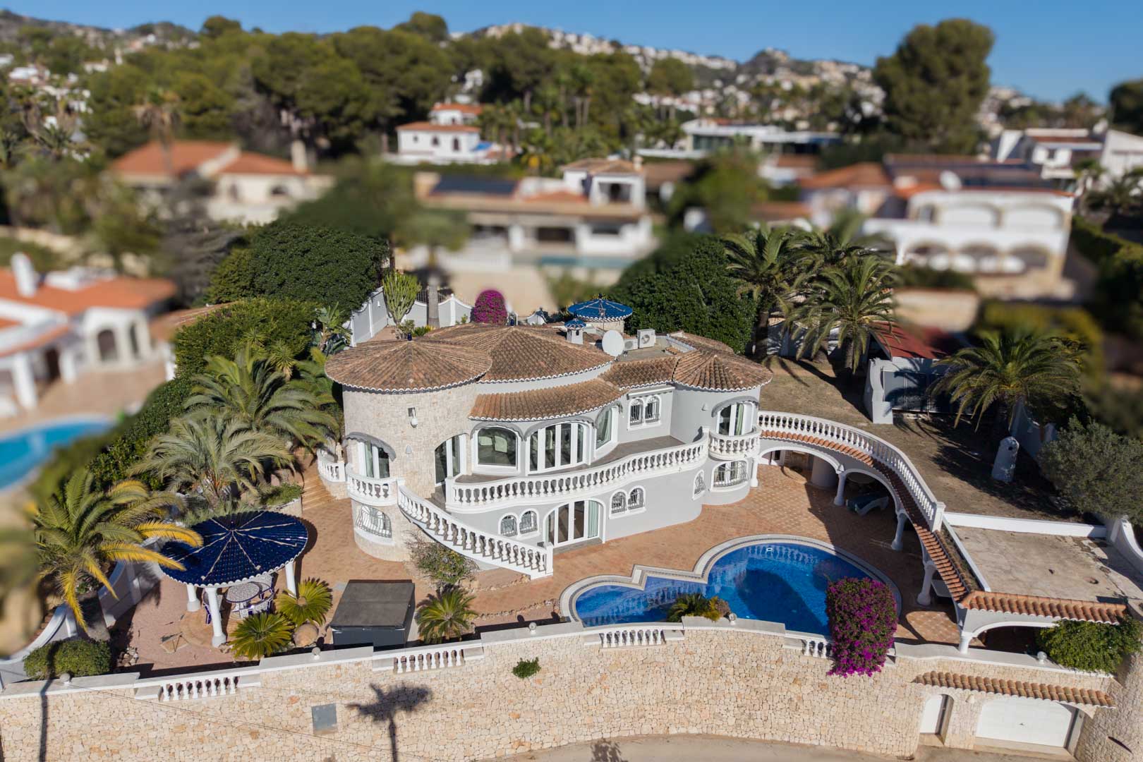 Luchtfoto van een mediterrane villa met natuurstenen toren, niervormig zwembad, gebogen witte balustrades en een tuin met palmbomen in een kustgebied.
