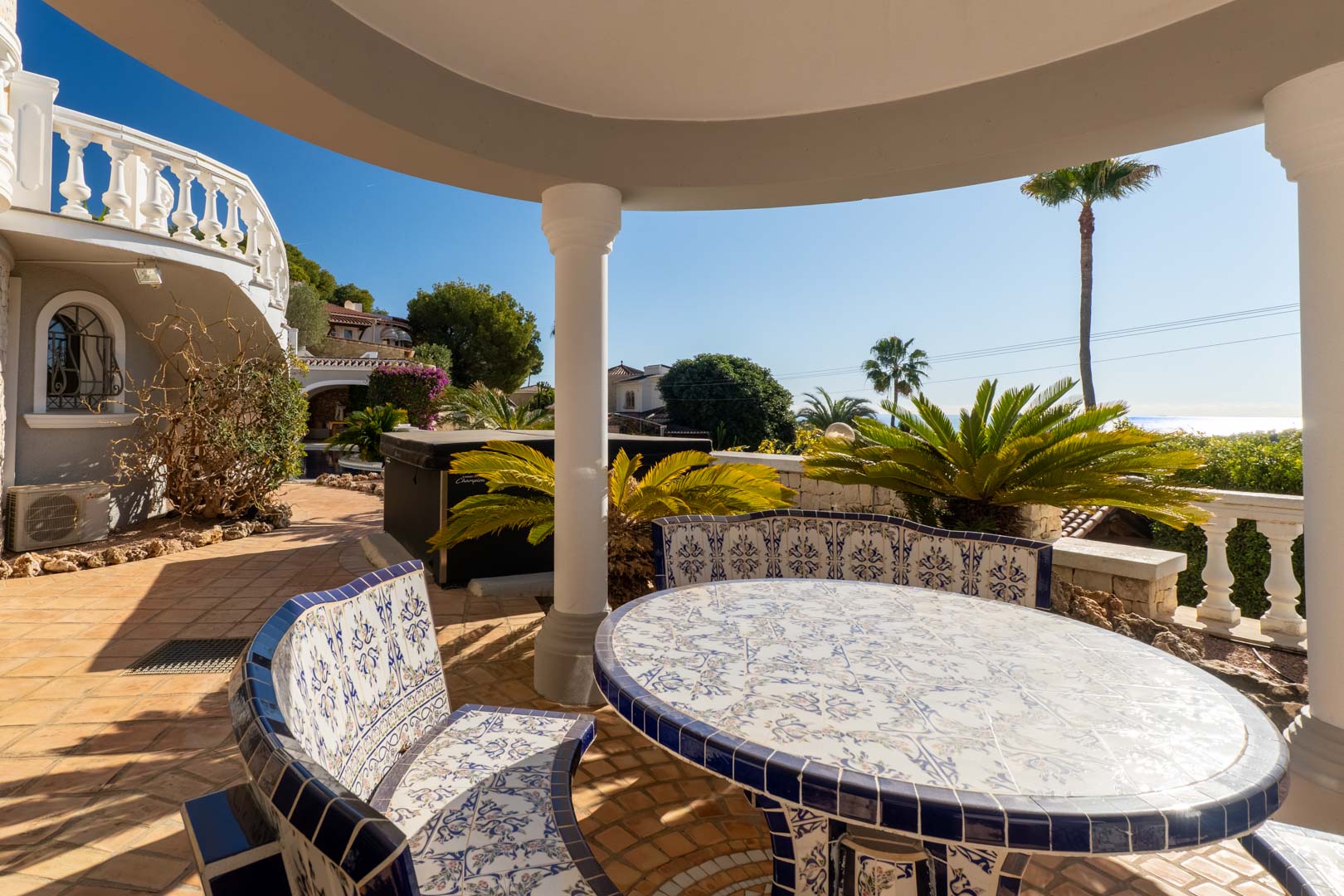 Overdekte eethoek buiten met een ronde tafel en banken van traditioneel blauw-wit keramiek, met uitzicht op de mediterrane tuin en de zee.
