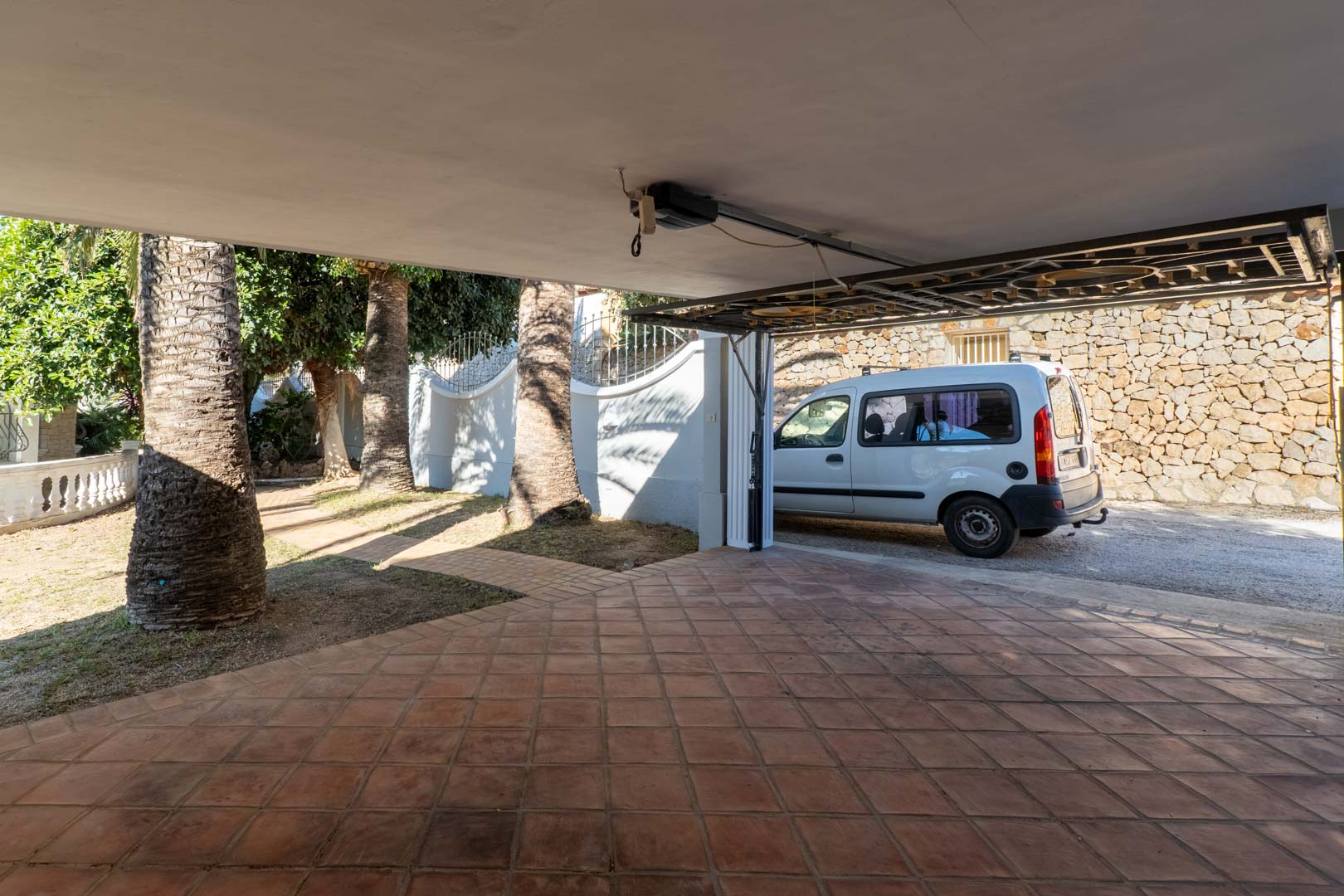 Overdekte garage met terracotta tegelvloer en automatische poort, grenzend aan een mediterrane tuin met palmbomen en natuurstenen muren.