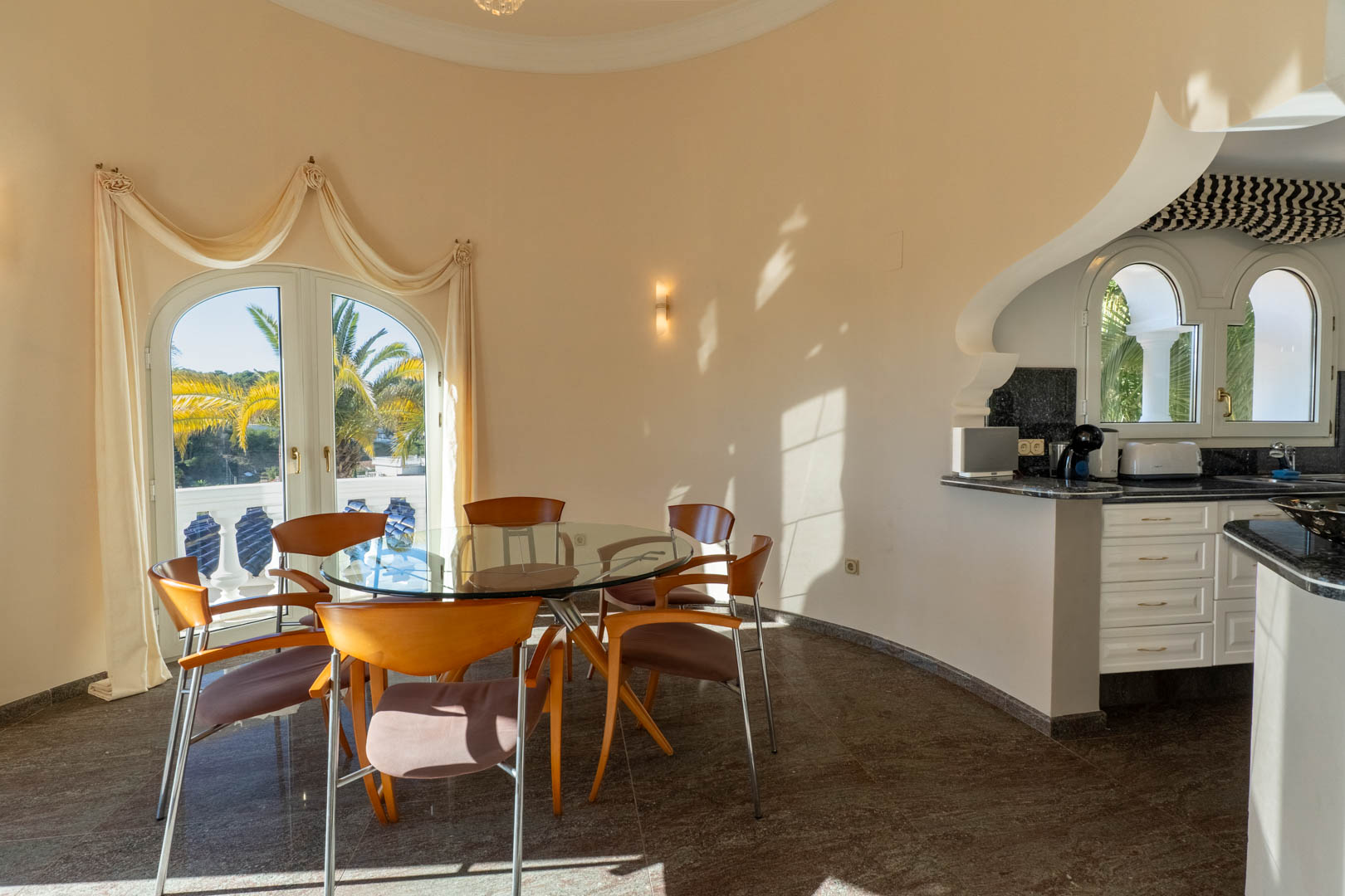Ronde eetkamer met glazen tafel en houten stoelen, verbonden met een keuken met granieten bladen en een boogdeur die uitkijkt op de mediterrane tuin.