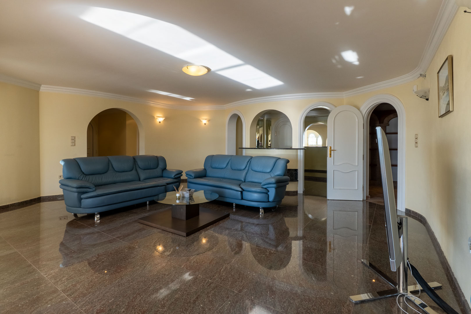 Ruime zithoek met glanzende marmeren vloer, blauwe lederen banken en architectonische bogen die leiden naar een bar en aangrenzende kamers.