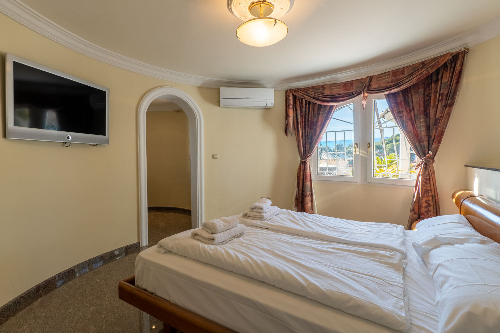 Slaapkamer met een gebogen wand, boogdeur en gepolijste stenen vloer. Voorzien van wand-tv, airconditioning en raam met uitzicht op de kustlijn.