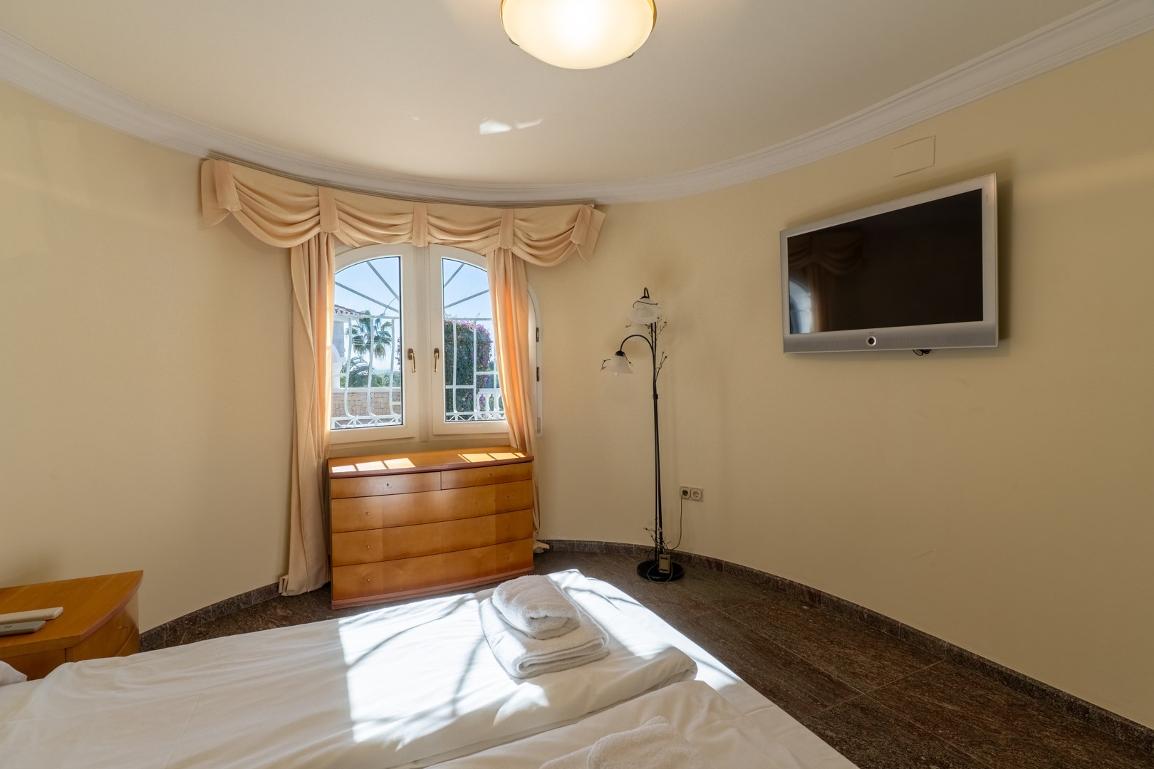 Slaapkamer met een gebogen wand, donkere granieten vloer en een boograam. Voorzien van een houten ladekast, wand-tv en veel natuurlijk lichtinval.