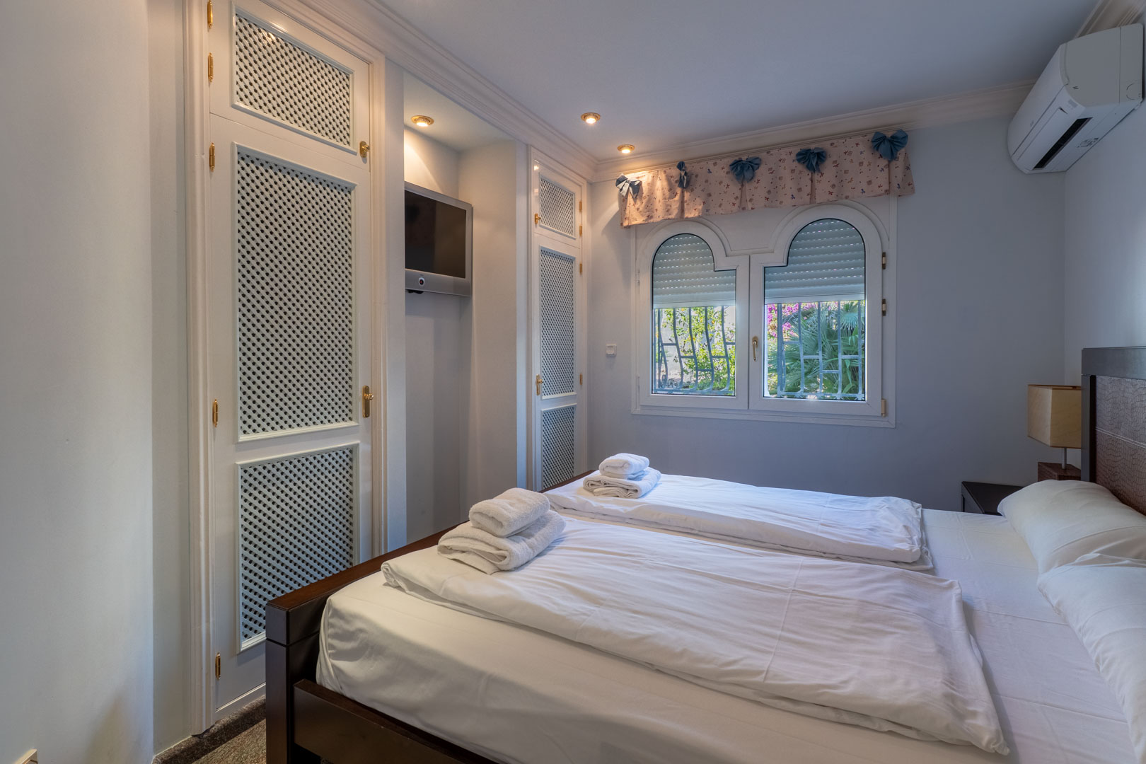 Slaapkamer met witte inbouwkasten met vlechtwerk, een geïntegreerde tv-nis en dubbele boogvensters met zicht op de tuin.