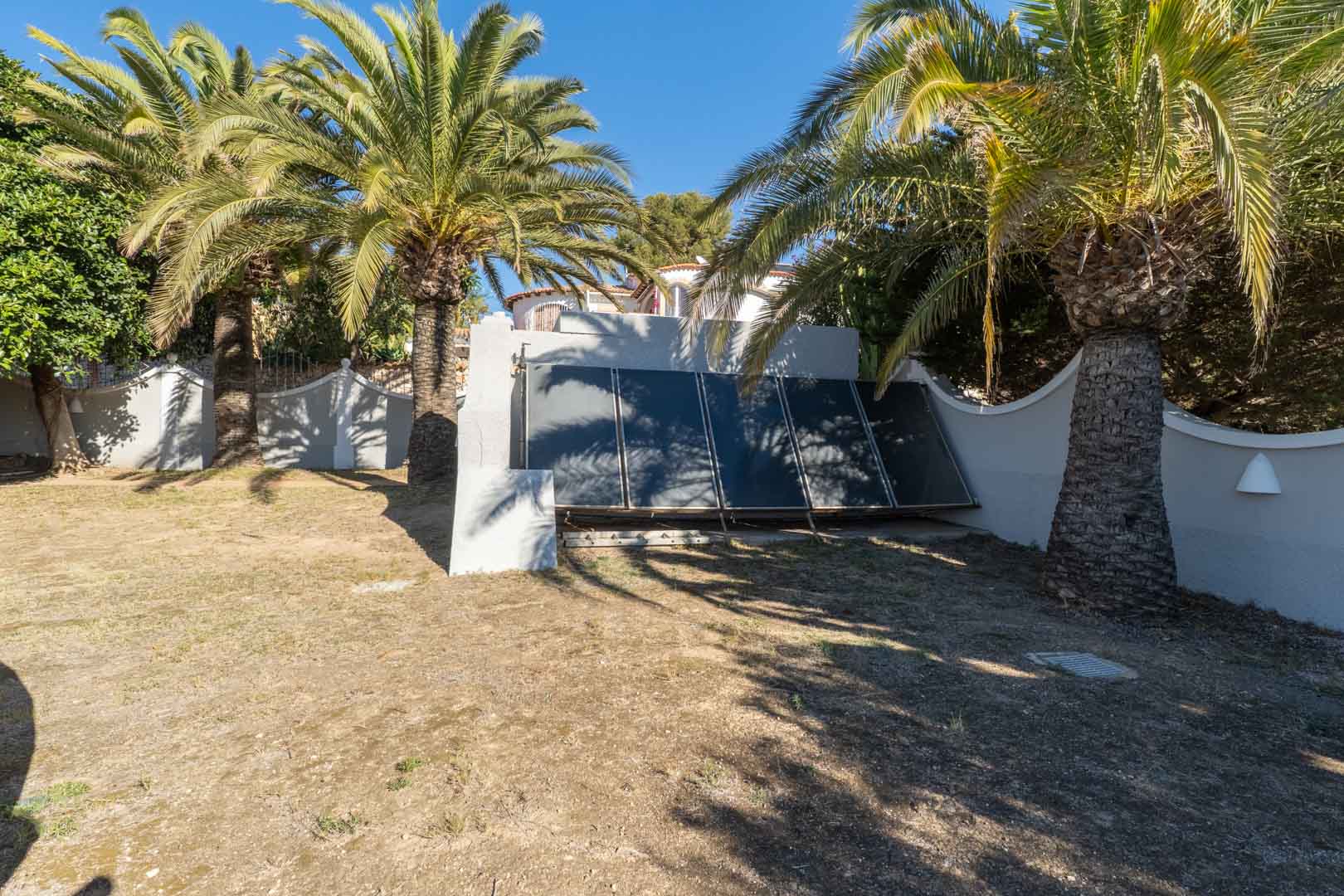 Tuinzone met op de grond gemonteerde thermische zonnepanelen, omgeven door volwassen palmbomen en witte muren in direct zonlicht.