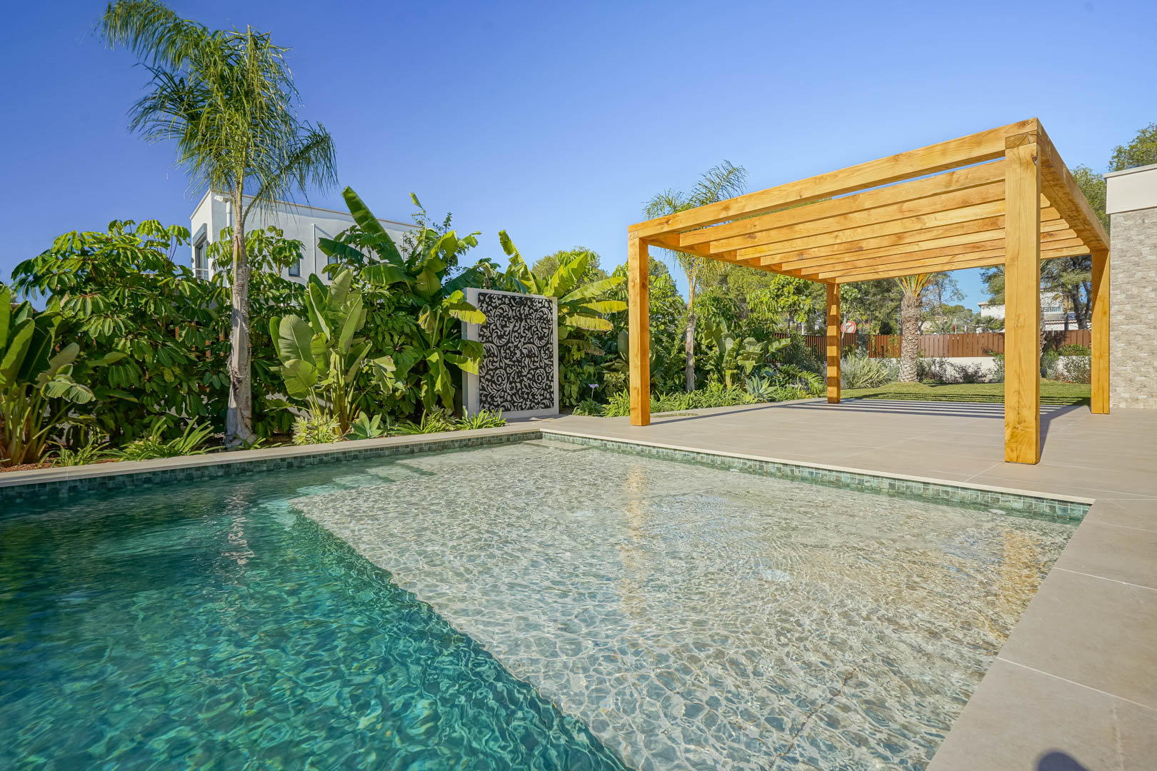 Bali-stijl zoutwaterzwembad met ondiep gedeelte, omgeven door een groot terras met een houten iroko pergola en een weelderige mediterrane tuin.