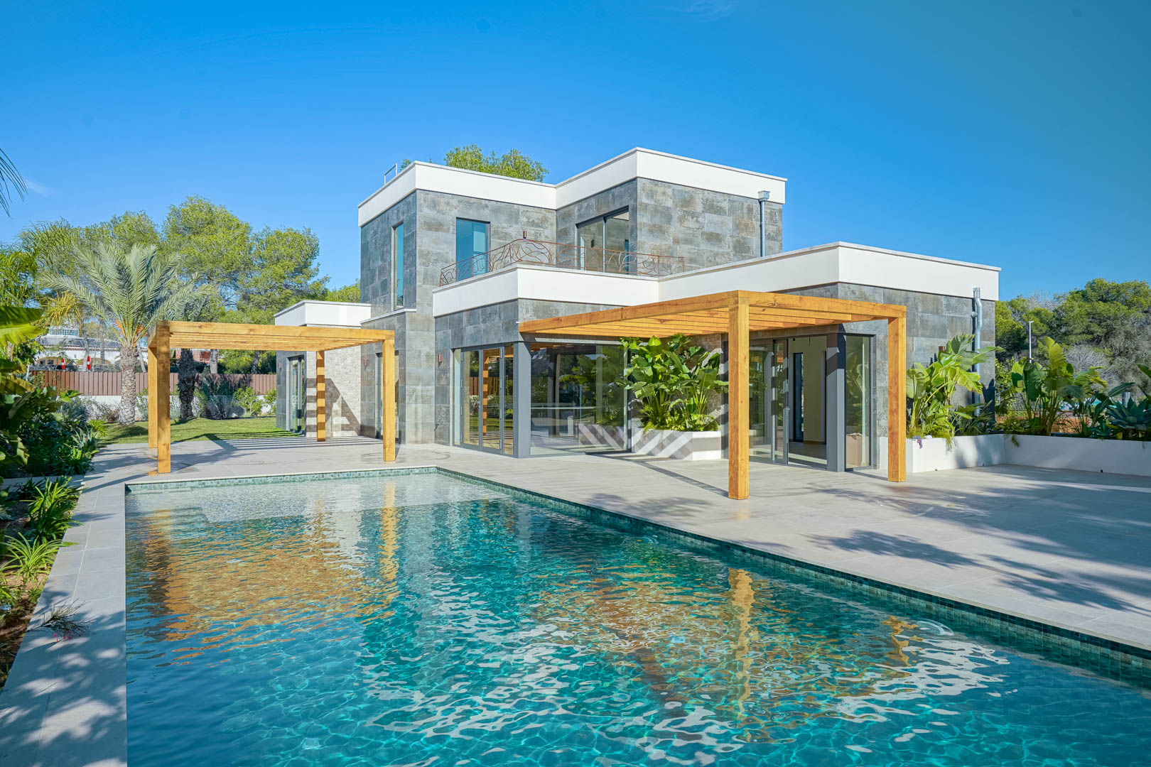 Buitenaanzicht van de villa met zwembad in Bali-stijl, ruime terrassen en pergola's van irokohout tegen een moderne gevel met natuurstenen afwerking.