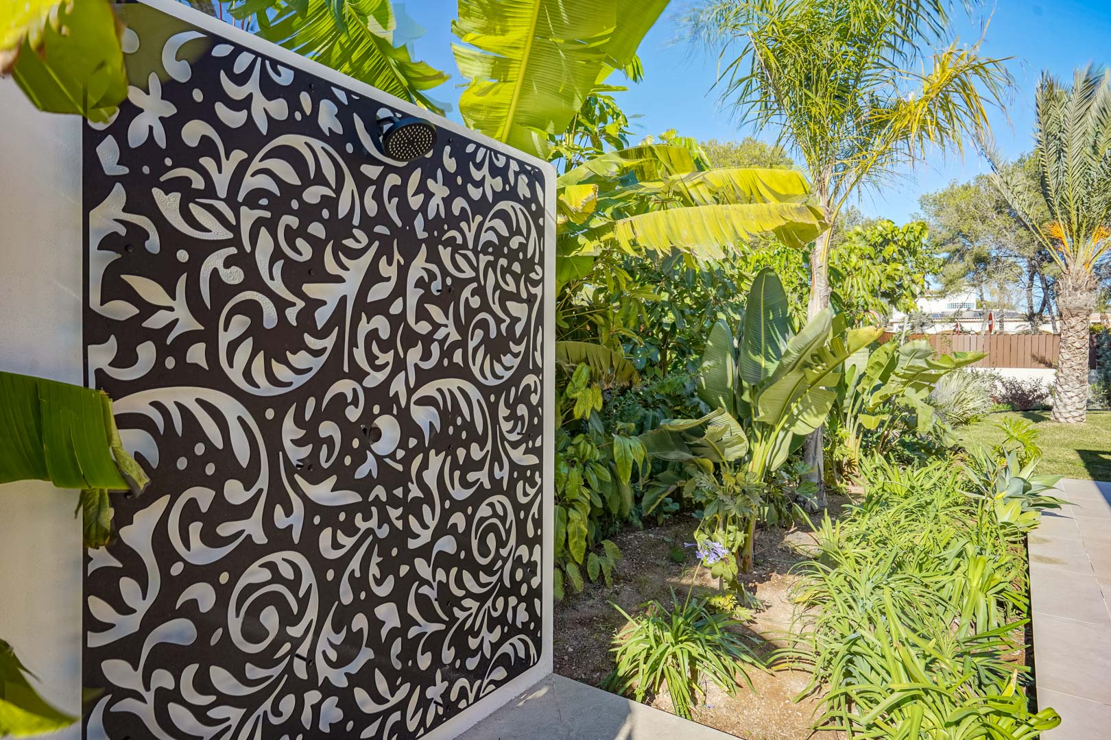 Buitendouche met een decoratief lasergesneden wandpaneel en zwarte kranen, omgeven door een weelderige mediterrane tuin met bananenbomen.