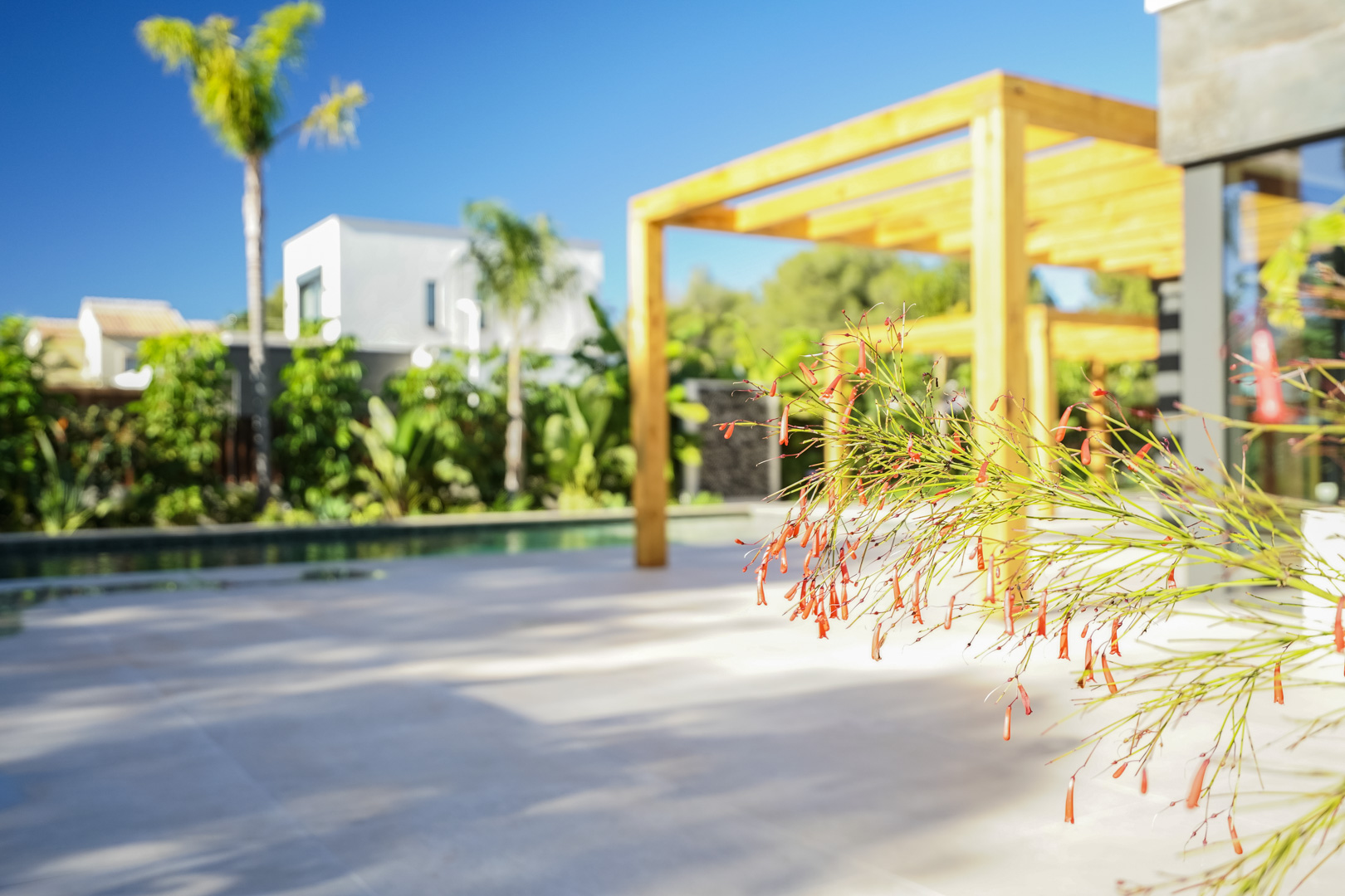 Buitenterras met een houten iroko pergola en uitzicht op de mediterrane tuin. Detail van de beplanting bij het zwembad en de lichte tegelvloer.