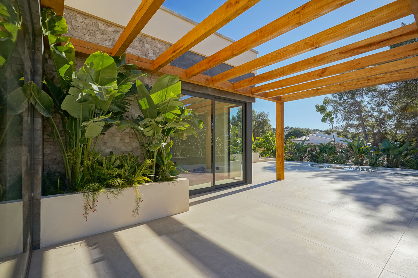 Buitenterras met een pergola van irokohout, grootformaat tegels en een ingebouwde plantenbak met tropische beplanting bij de natuurstenen gevel.
