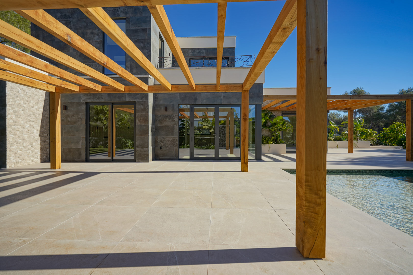 Buitenterras met iroko houten pergola en lichte tegels, gelegen naast het zwembad en een moderne gevel met grote glazen deuren.