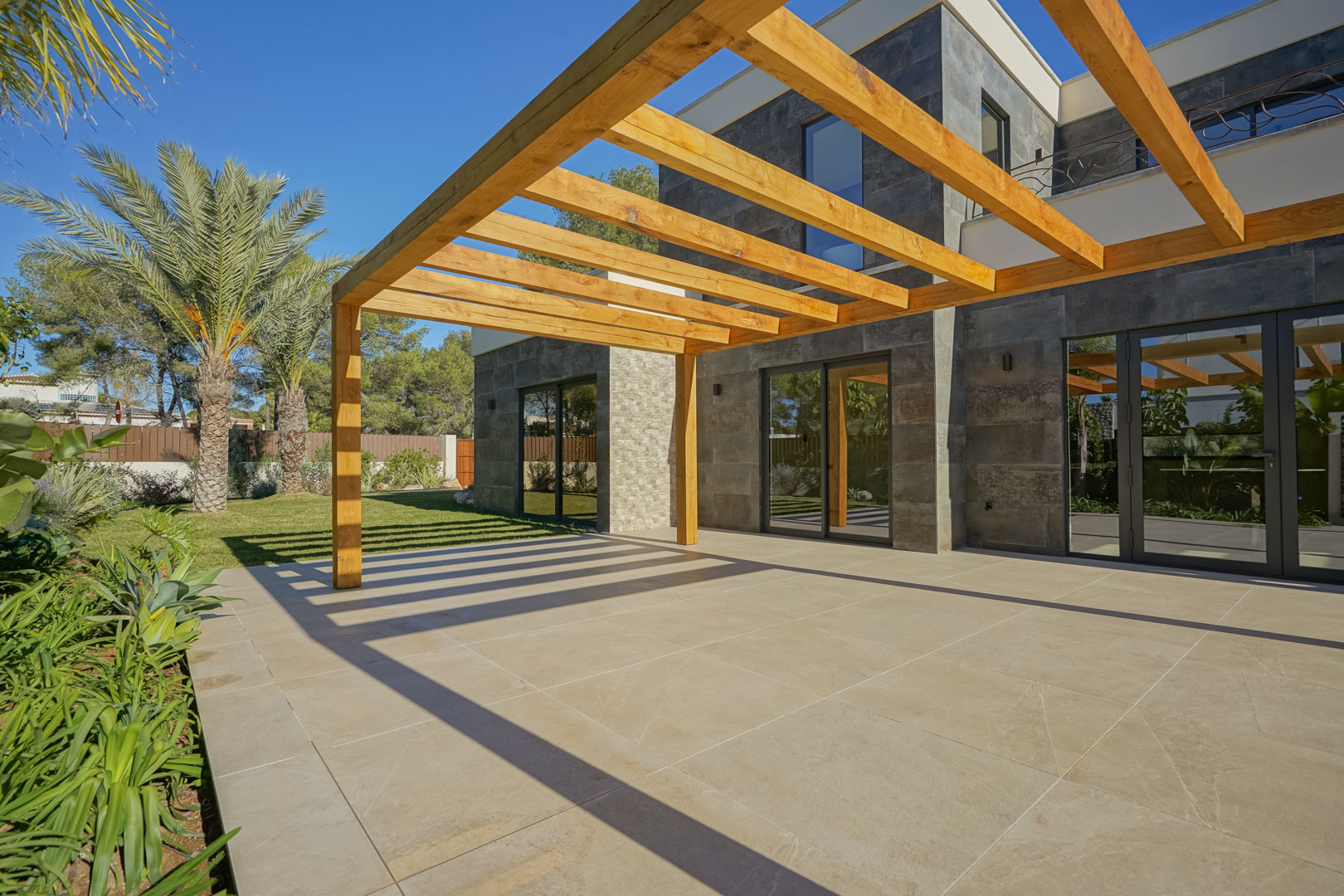 Buitenterras voorzien van een iroko houten pergola en grootformaat tegels, grenzend aan een natuurstenen gevel en een aangelegde mediterrane tuin.