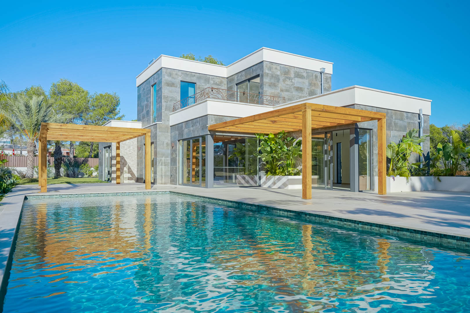 Exterieur van de villa met een turquoise mozaïekzwembad, iroko houten pergola's en een natuurstenen gevel met grote glazen puien.
