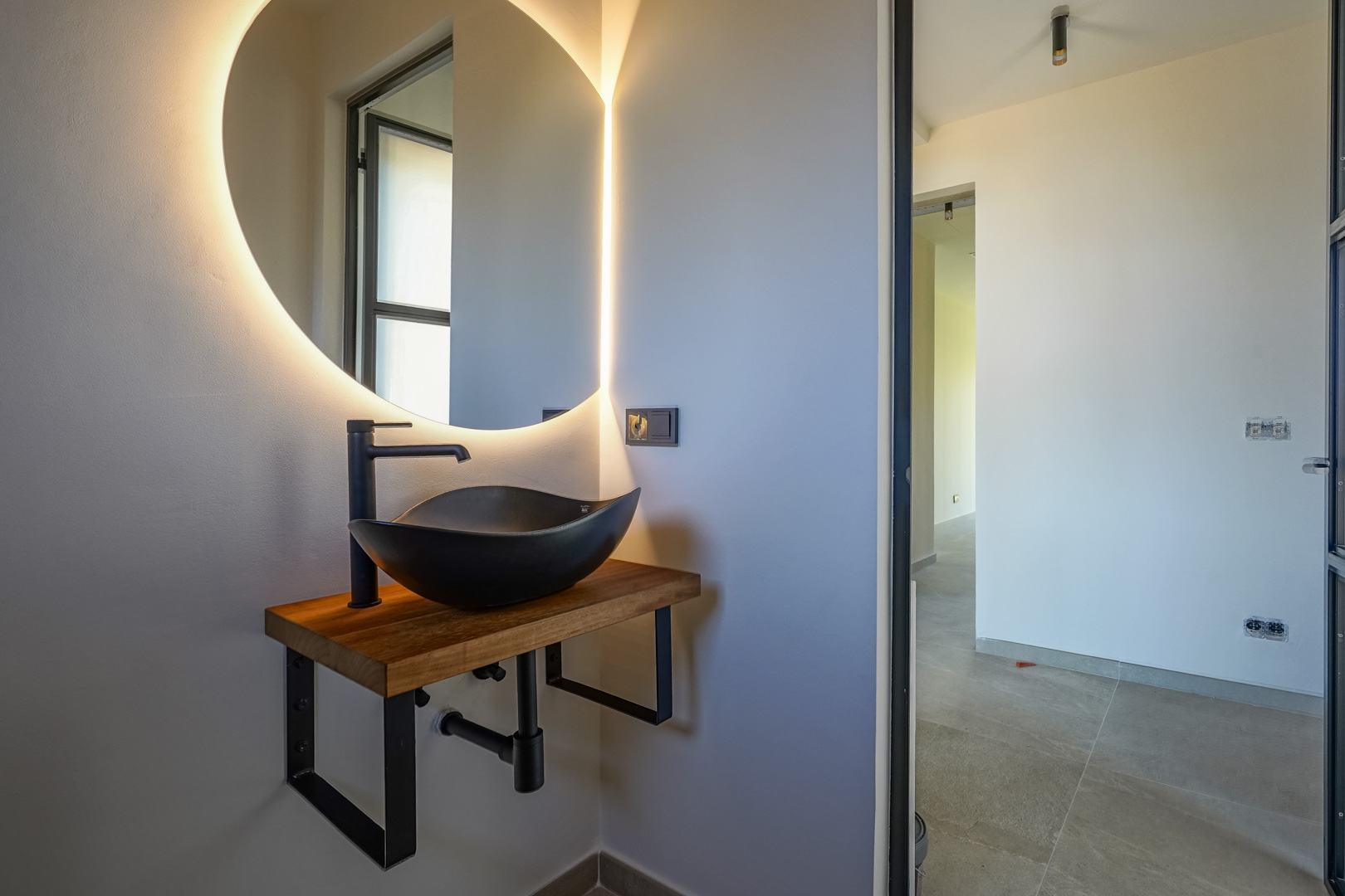Gastentoilet met een zwarte opzetwastafel op een massief houten blad, voorzien van een ronde spiegel met geïntegreerde led-achtergrondverlichting.