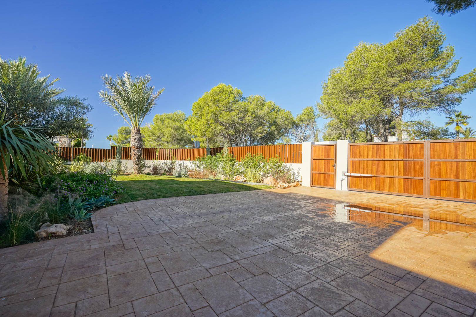 Hoofdingang met gemotoriseerde houten poort, bestrate oprit en een aangelegde mediterrane tuin met palmbomen en houten privacy-omheining.