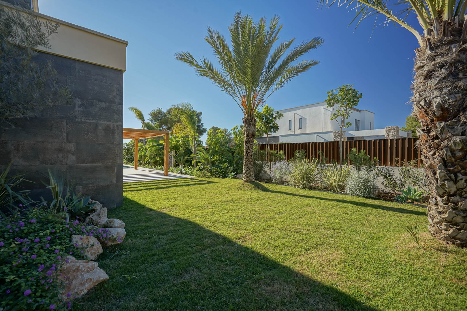 Mediterrane tuin met natuurlijk gazon, palmbomen en inheemse beplanting naast een donkere stenen gevel en een houten iroko pergola.
