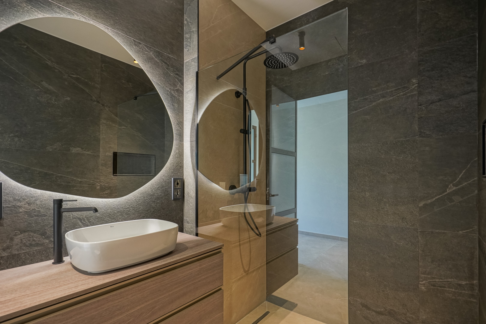 Moderne badkamer met donkere tegels in steenlook, houten wastafelmeubel met opbouwkom en een ronde verlichte spiegel naast een inloopdouche.