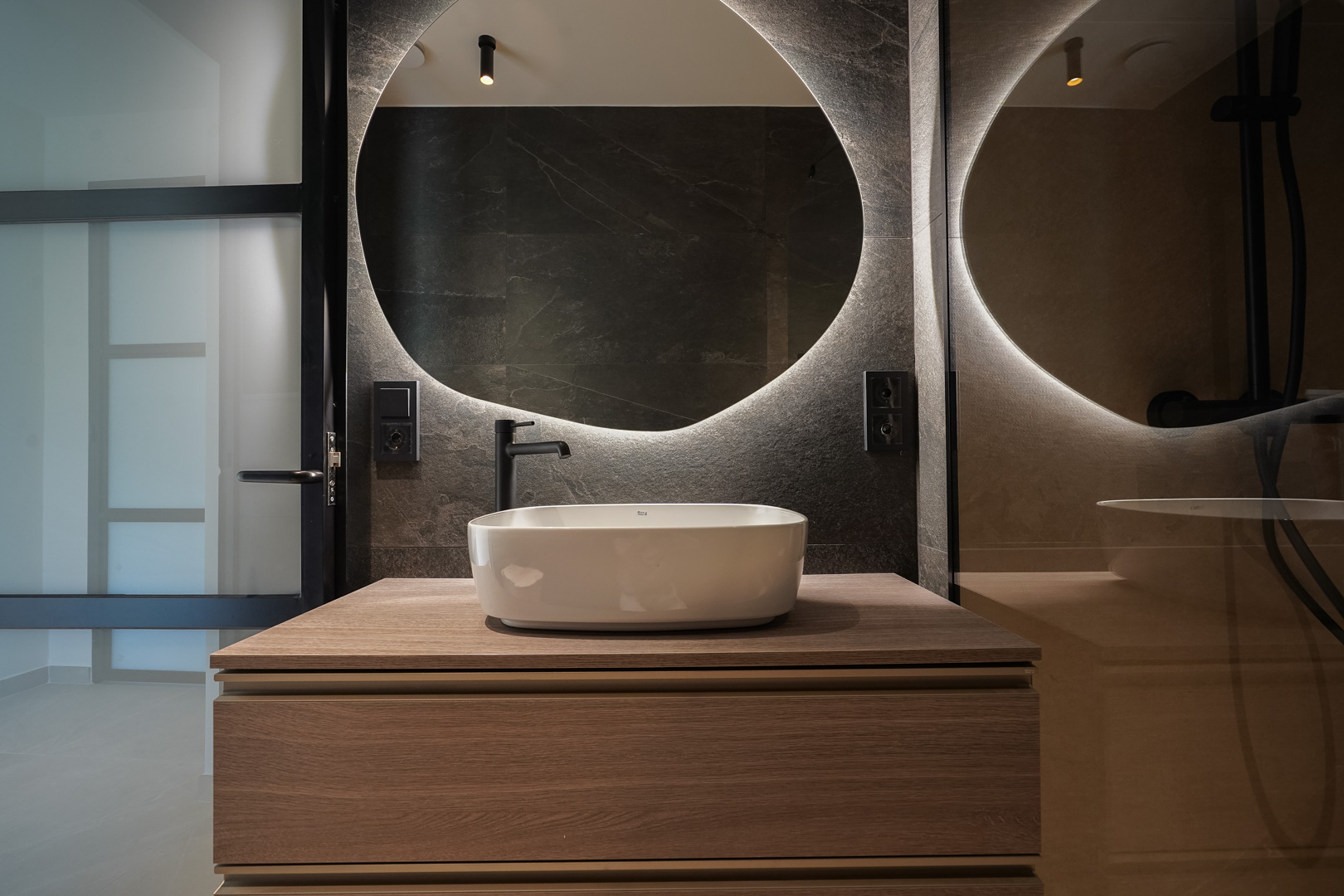 Moderne badkamer met een opzetwastafel op een houten meubel, een ronde indirect verlichte spiegel en donkere wandtegels met matzwarte kranen.