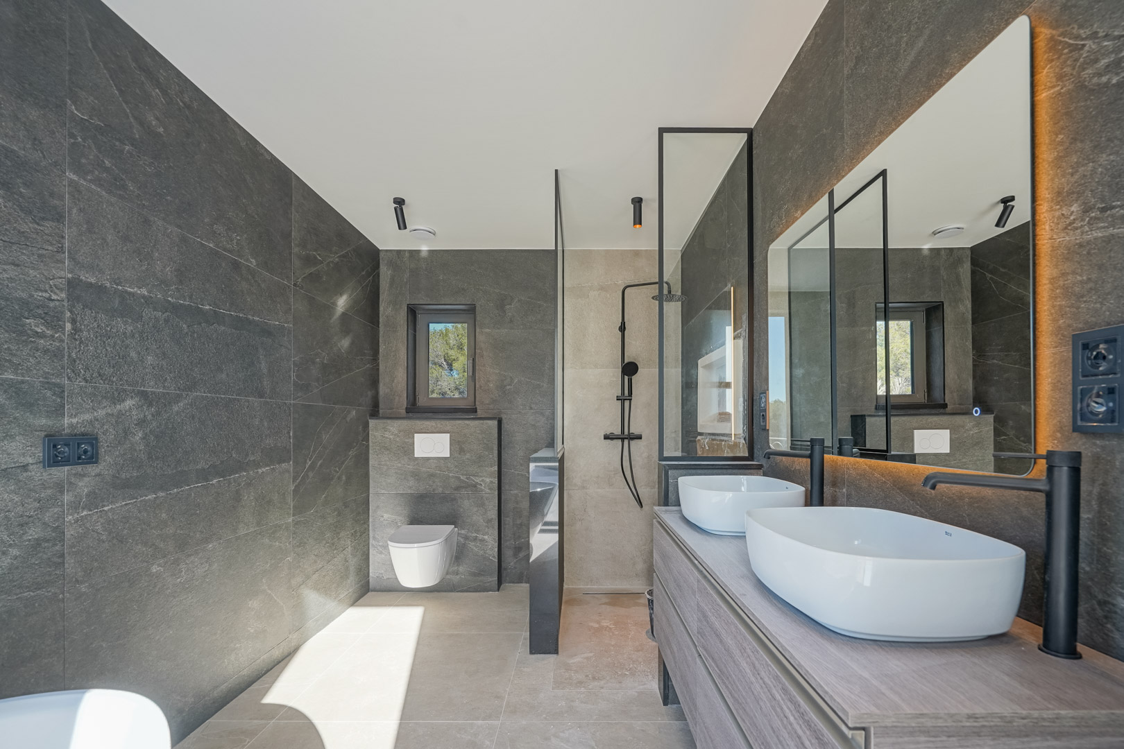 Moderne badkamer met grijze natuursteenlook tegels, dubbele opzetwastafels op een houten meubel, matzwarte kranen en een inloopdouche met glazen wand.