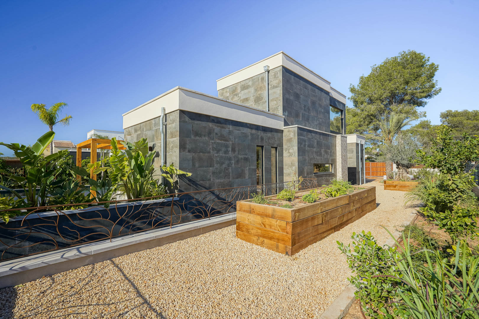 Moderne gevel met donkere natuursteen, verhoogde houten plantenbakken en grindpaden in een aangelegde mediterrane tuin.