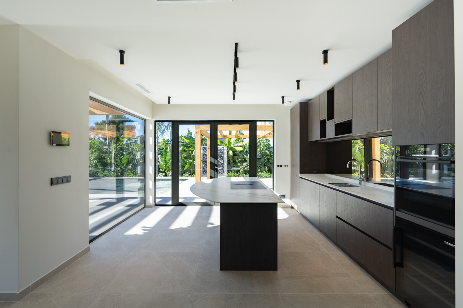 Moderne keuken met kookeiland, donkerhouten kasten en stenen werkbladen. Grote glazen puien bieden direct zicht op de tuin met zwembad en houten pergola.