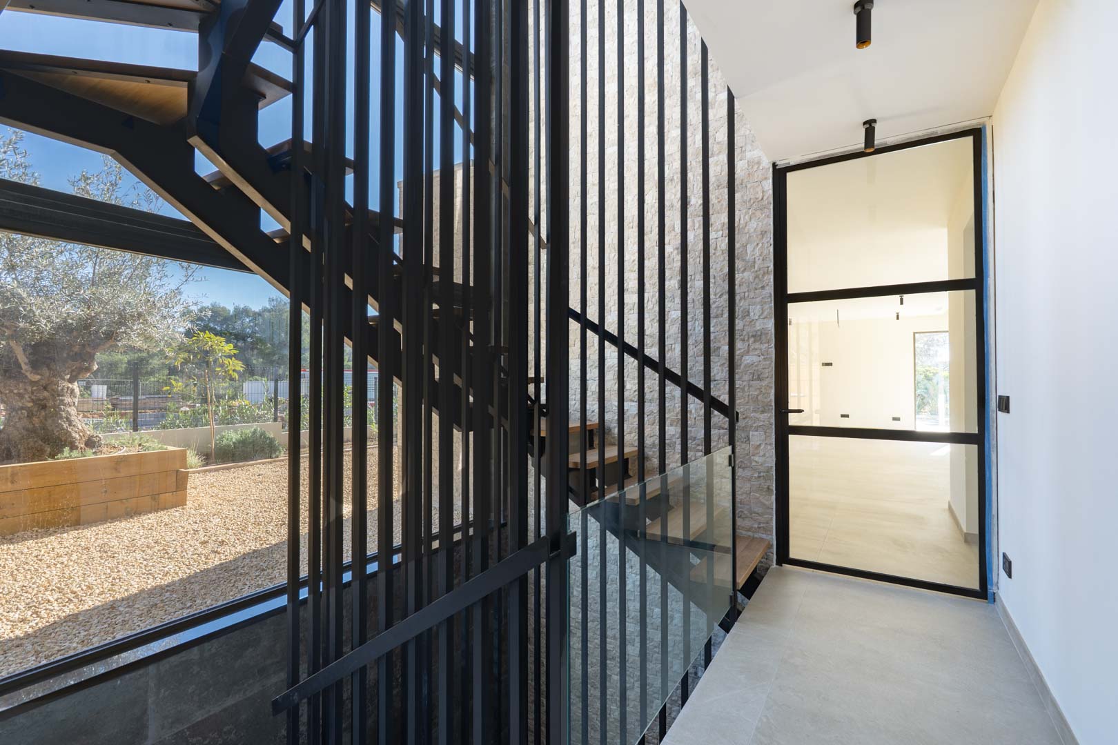 Moderne trap met houten treden, zwart metalen spijlen en een natuurstenen wand, verlicht door grote raampartijen met uitzicht op de karakteristieke olijfboom.