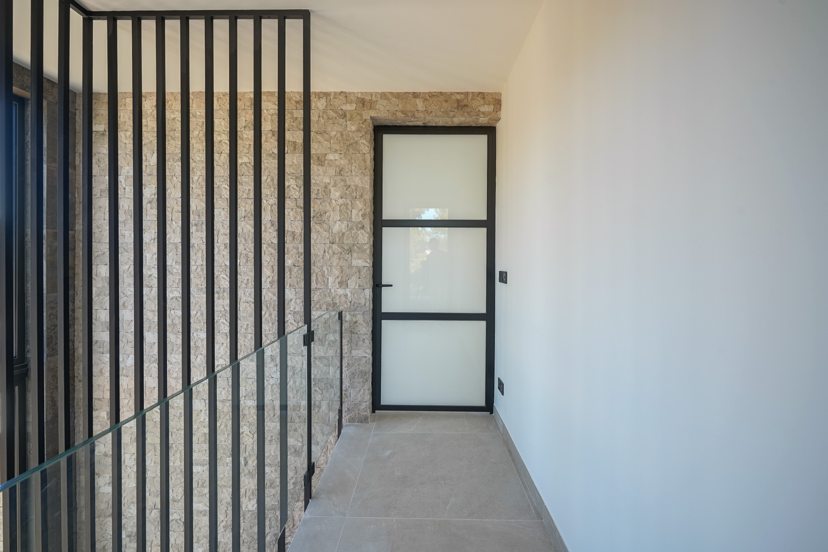 Overloop op de bovenverdieping met een natuurstenen wand, glazen balustrade met zwarte lamellen en een industriële deur met matglas.