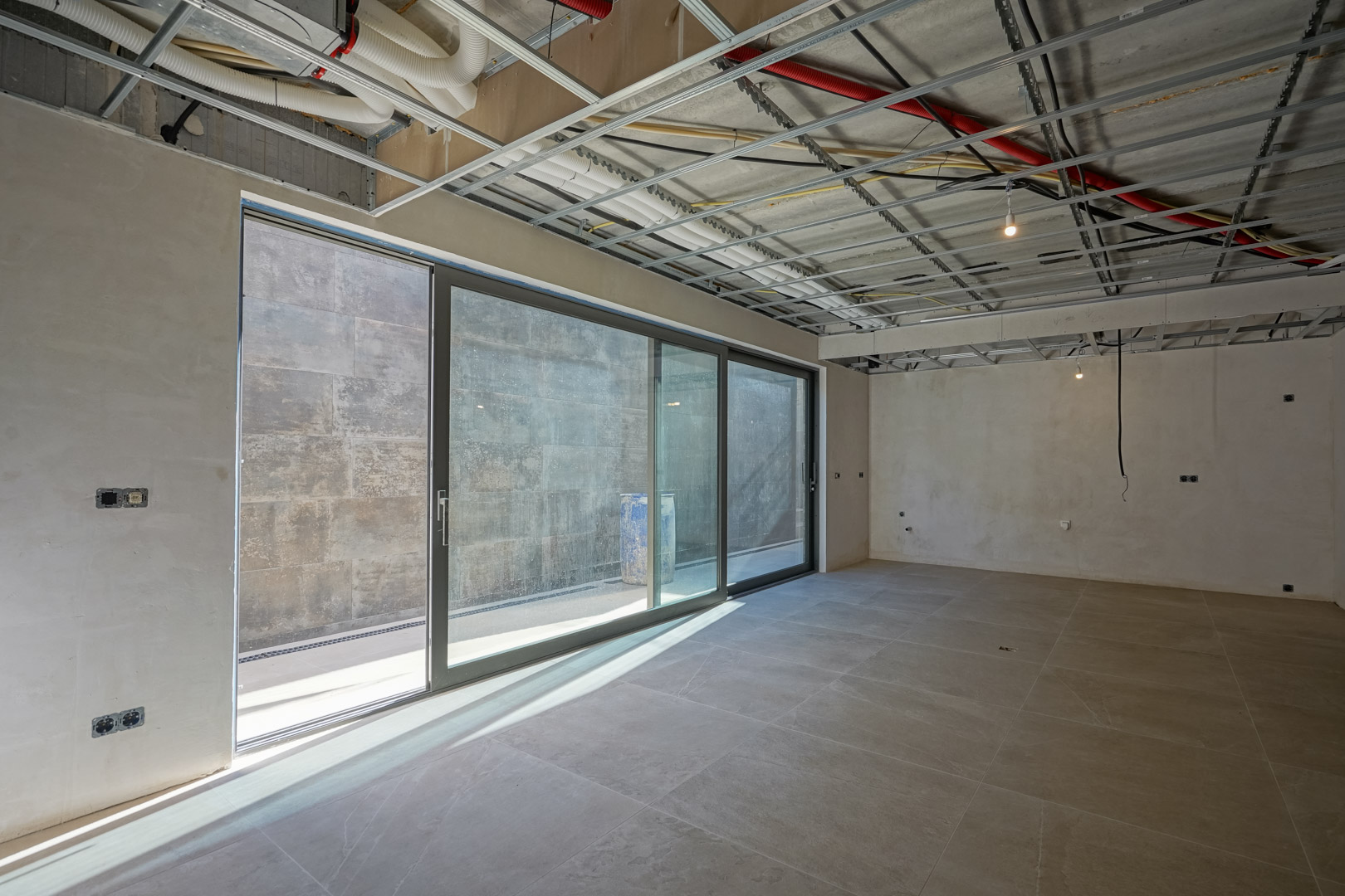 Souterrain in afbouwfase met grote glazen schuifdeuren, grootformaat vloertegels en zichtbare technische installaties aan het plafond voor ventilatie en elektra.