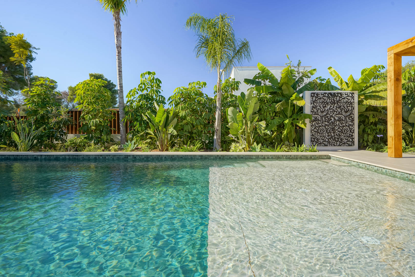 Zoutwaterzwembad in Bali-stijl met ondiep gedeelte, omgeven door een weelderige mediterrane tuin met tropische beplanting en een decoratief paneel.