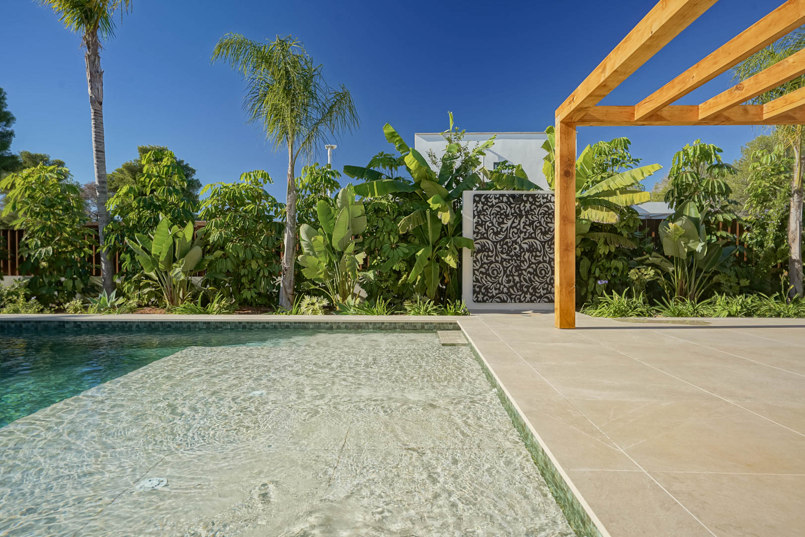 Zoutwaterzwembad in Bali-stijl met ondiep loungeterras, omringd door een ruim zonneterras, een iroko houten pergola en een weelderige tropische tuin.