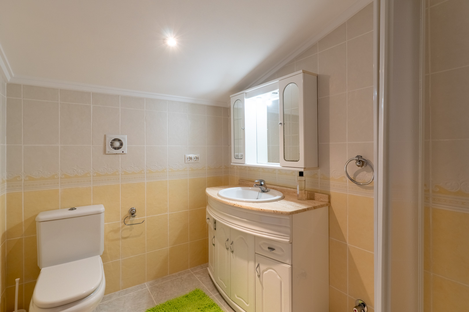 Badkamer met crème en gele tegels, een gebogen wastafelmeubel met marmeren blad en een spiegelkast met geïntegreerde verlichting.