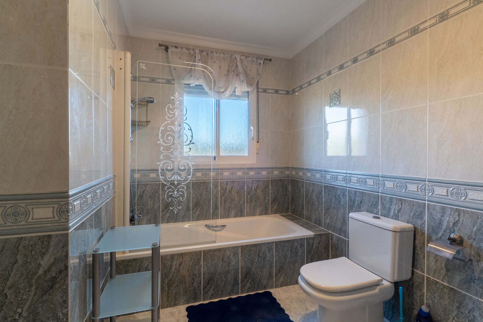Badkamer met ingebouwd ligbad, glazen douchescherm met decoratief patroon en tweekleurige betegeling. Voorzien van een raam voor natuurlijk licht.