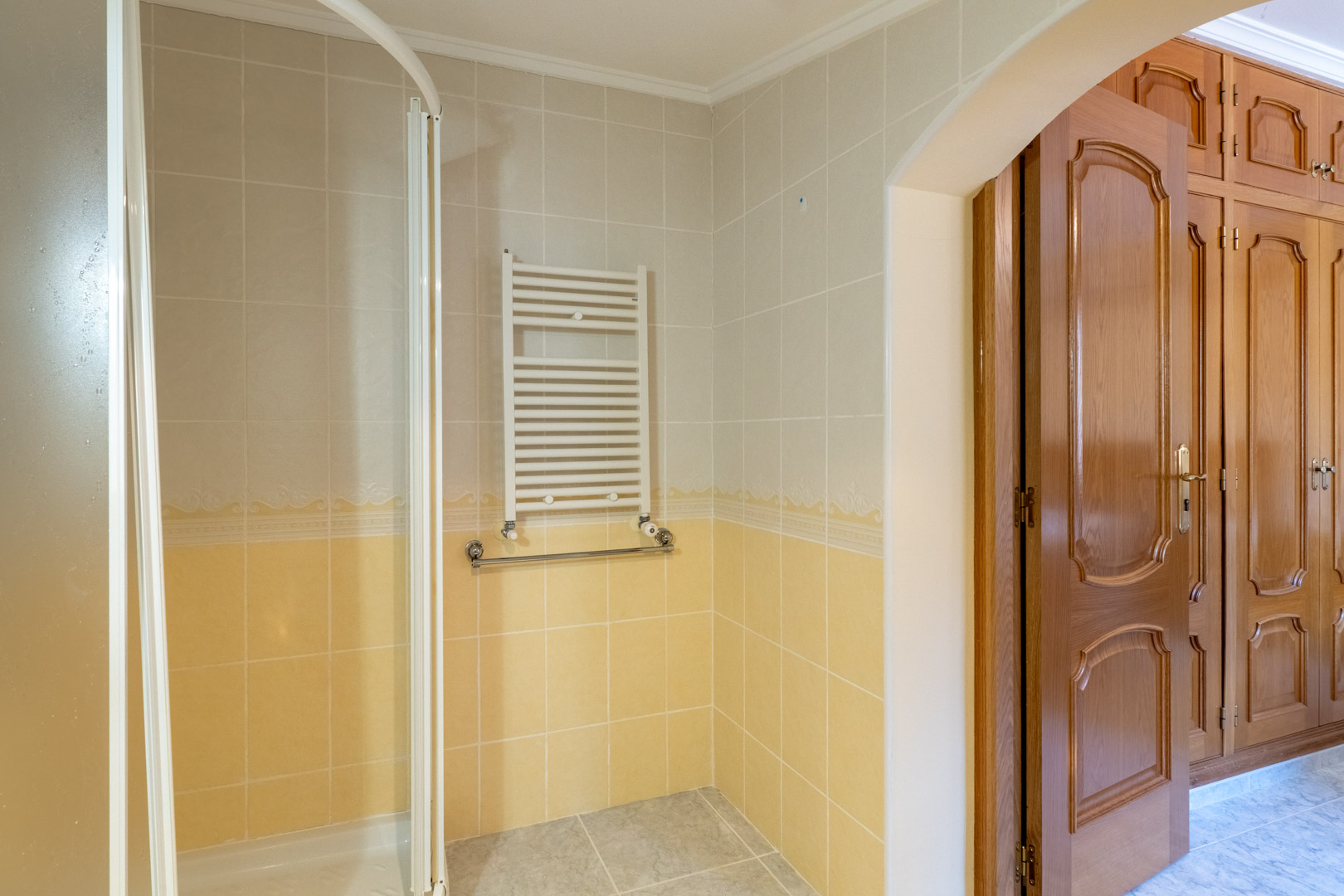 Badkamer met tweekleurige tegels, handdoekradiator en douche, via een boog verbonden met de slaapkamer met klassieke houten inbouwkasten.