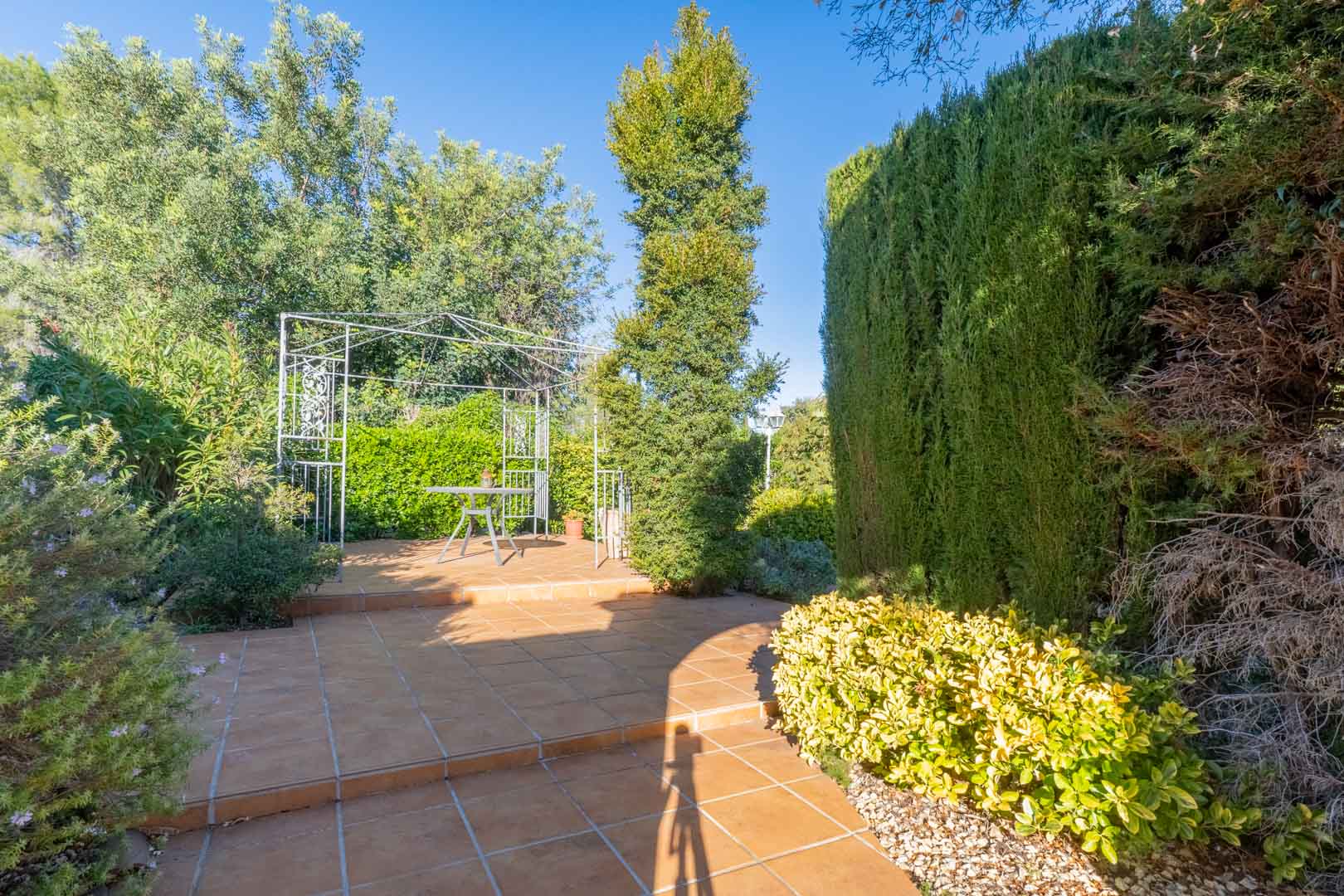 Buitenterras met keramische tegels, een witte metalen pergola en volwassen mediterrane beplanting die zorgt voor natuurlijke privacy.