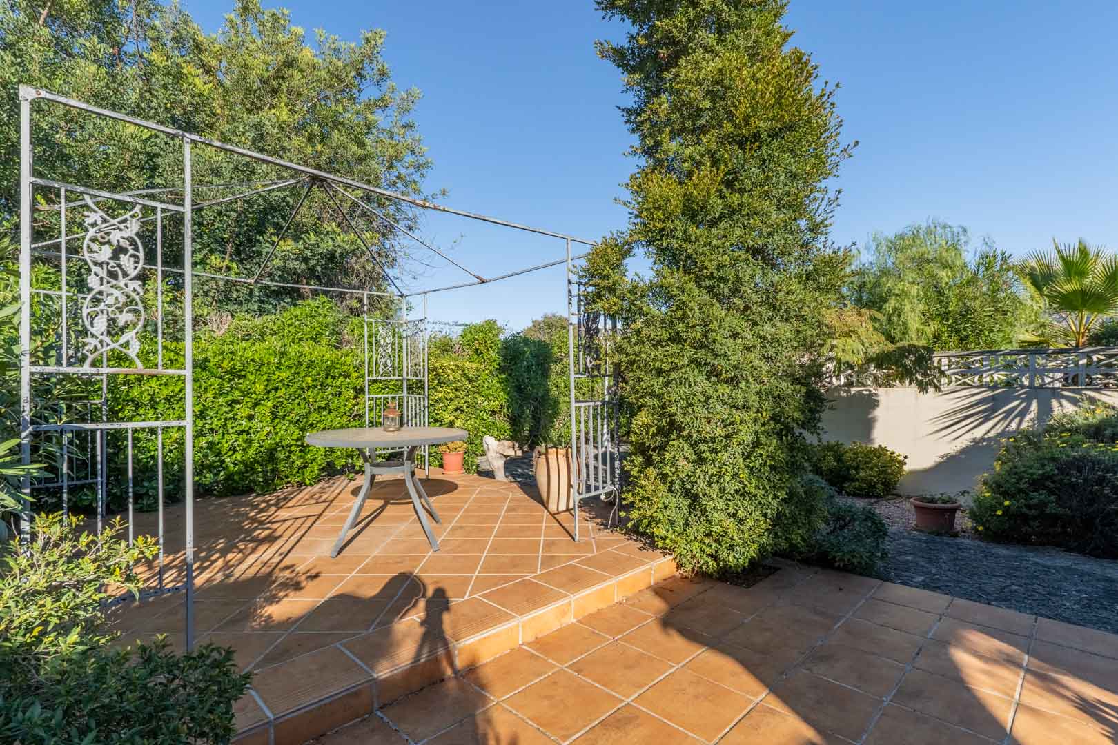 Buitenterras met terracotta tegels, een decoratieve metalen pergola en een ronde tafel, omgeven door volgroeide mediterrane beplanting en hagen.