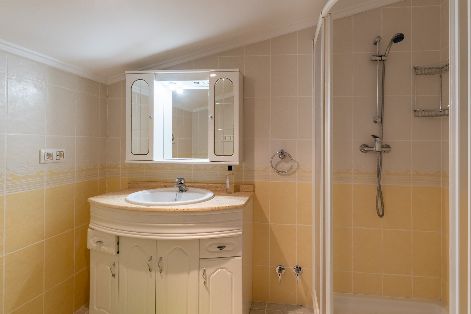 Functionele badkamer met een marmeren wastafelmeubel, witte opbergkast en douche, afgewerkt met crème- en geeltinten tegels en een spiegelkast.