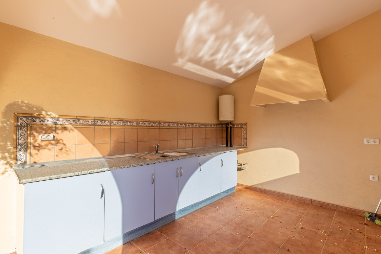 Functionele buitenkeuken met granieten werkblad, lichtblauwe onderkasten en traditionele tegelwand. Voorzien van een roestvrijstalen spoelbak en een grote afzuigkap.