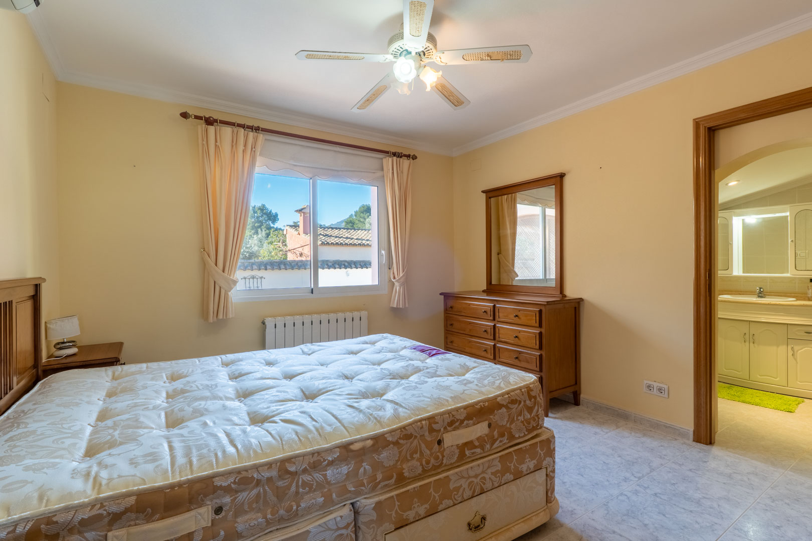 Hoofdslaapkamer met houten meubilair, plafondventilator en een aangrenzende badkamer. De kamer is voorzien van een radiator en een raam met natuurlijk licht.