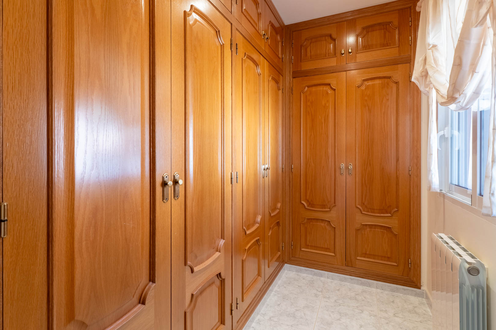 Kleedkamer met ruime houten inbouwkasten met klassieke panelen, een tegelvloer en natuurlijke lichtinval via het zijraam.