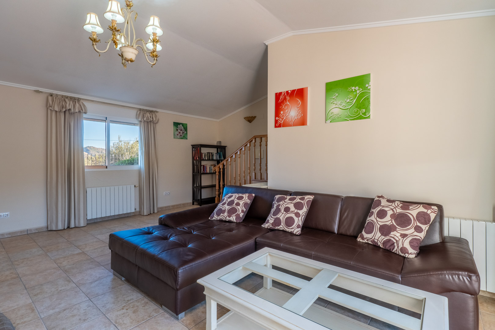 Lichte woonkamer met bruin lederen hoekbank, witte salontafel en tegelvloer. Voorzien van een houten trap, radiator en een klassieke kroonluchter aan het plafond.