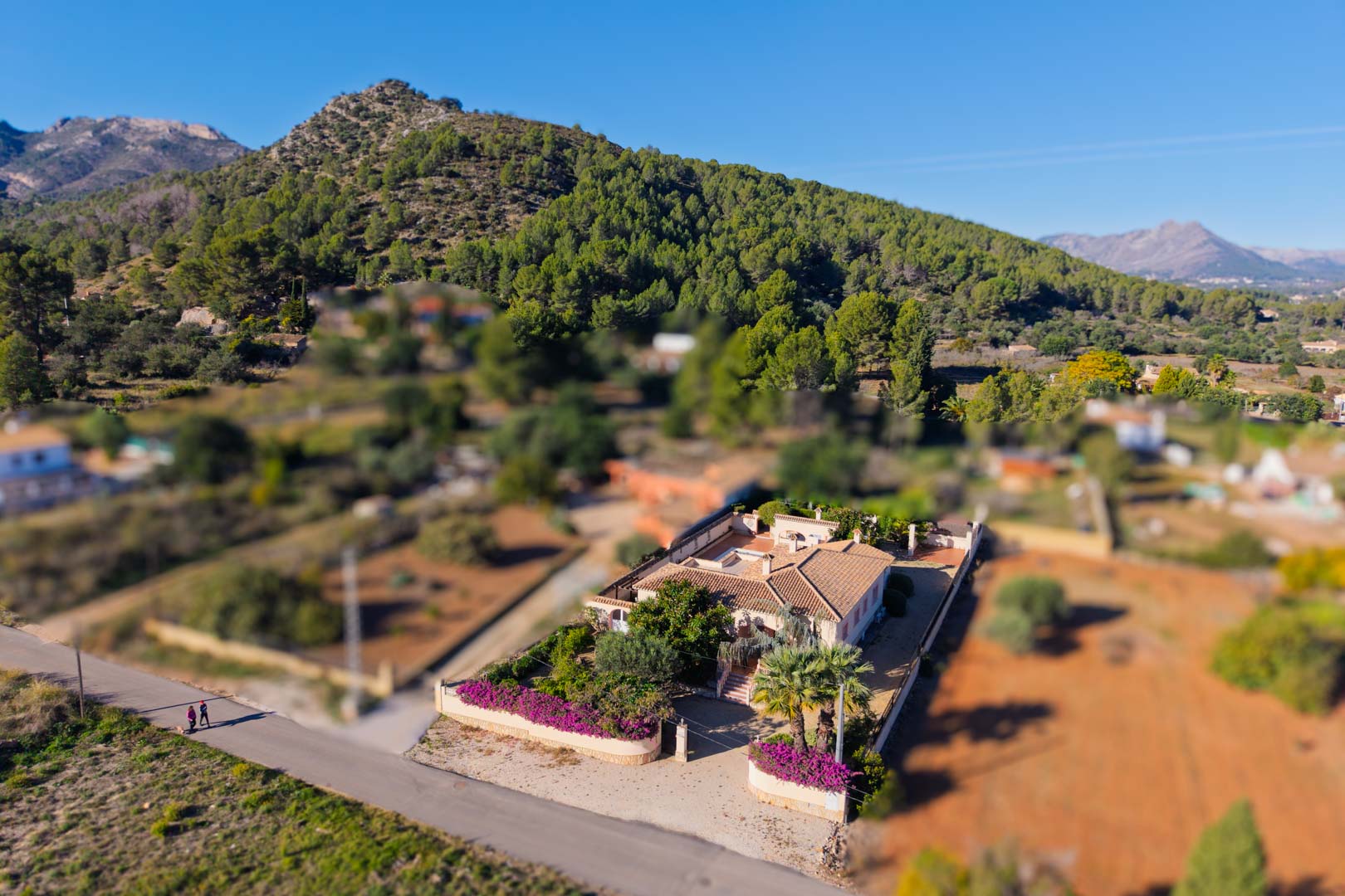 Luchtfoto van de mediterrane villa op een vlak perceel, met een traditioneel pannendak en tuinen vol bougainvillea aan de voet van de bergen.