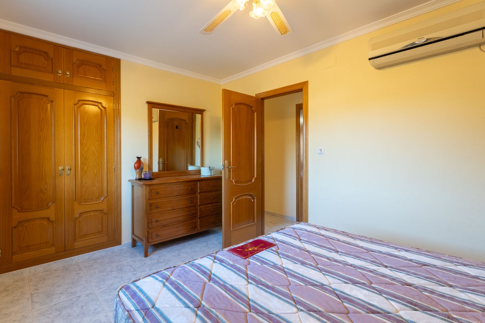 Slaapkamer met ingebouwde houten kasten, bijpassende ladekast met spiegel, airconditioning en plafondventilator op een lichte tegelvloer.