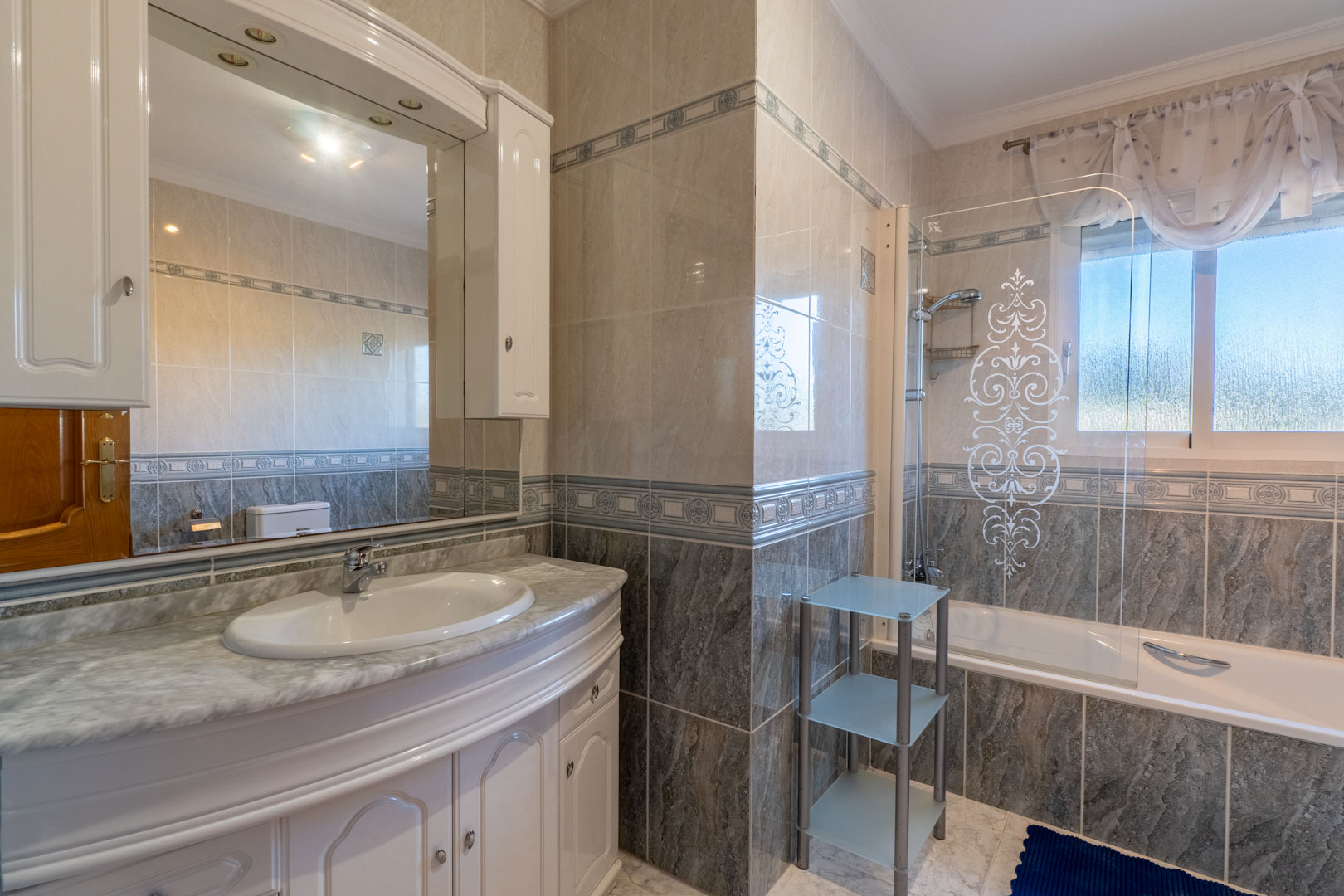 Volledige badkamer met marmeren wastafelblad, witte kasten en ligbad met een gedecoreerd glazen scherm. Betegelde wanden met sierranden en natuurlijk licht.